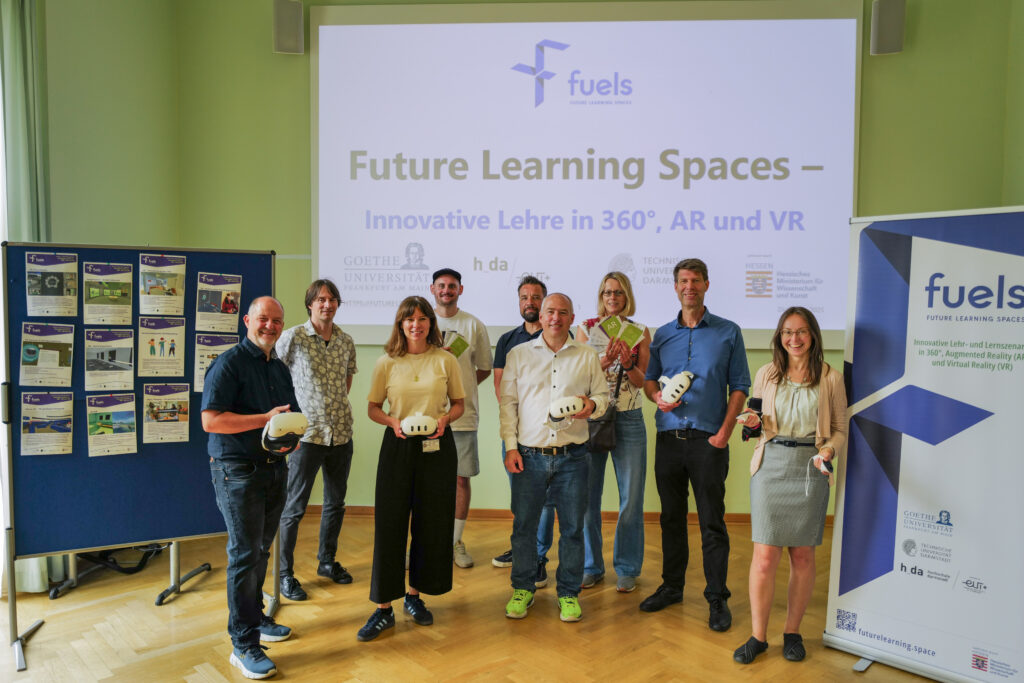 Gruppenfoto des fuels-Teams, mit VR-Brillen in der Hand. Im Hintergrund eine Projektion mit Schriftzug "Future Learning Spaces - innovative Lehre in 360°, AR und VR".